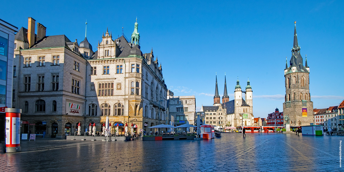 KI generiert: Das Bild zeigt den Marktplatz in Halle (Saale) mit einer beeindruckenden architektonischen Kulisse, bestehend aus historischen Gebäuden und einer Kirche mit hohen Türmen. Der Platz wirkt belebt und ist bei klarem Himmel fotografiert.