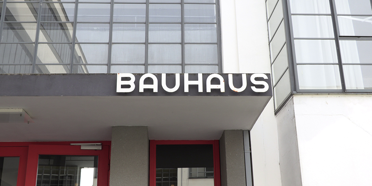 KI generiert: Das Bild zeigt den Eingang eines Gebäudes mit der Aufschrift "BAUHAUS" in markanten, modernen Buchstaben. Die Architektur ist nüchtern und funktional gestaltet, typisch für den Bauhaus-Stil.
