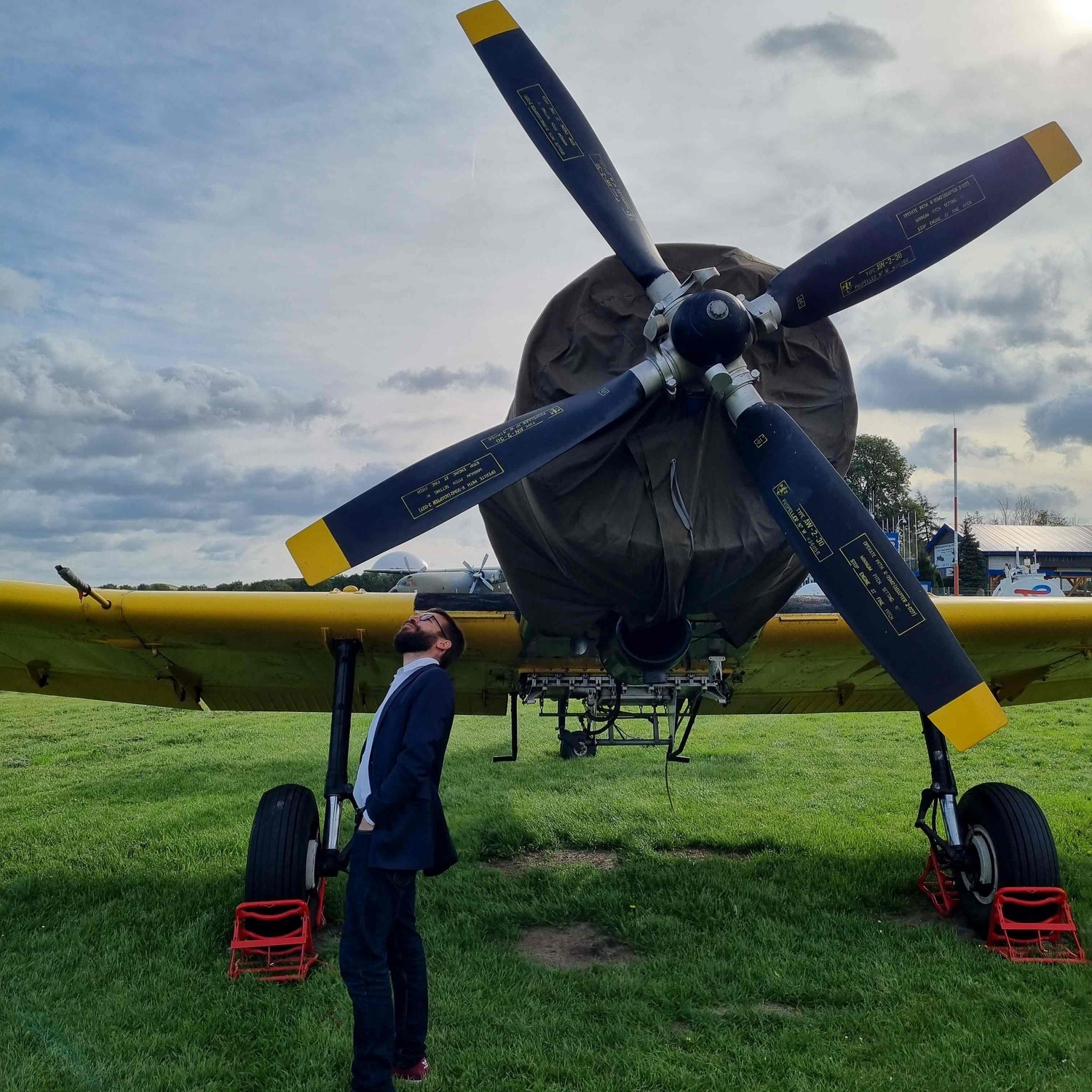 KI generiert: Das Bild zeigt einen großen Propeller eines Flugzeugs auf einem Feld, vor dem ein Mann steht und nach oben schaut. Das Flugzeug hat eine Abdeckung und steht auf einem grasbewachsenen Untergrund.