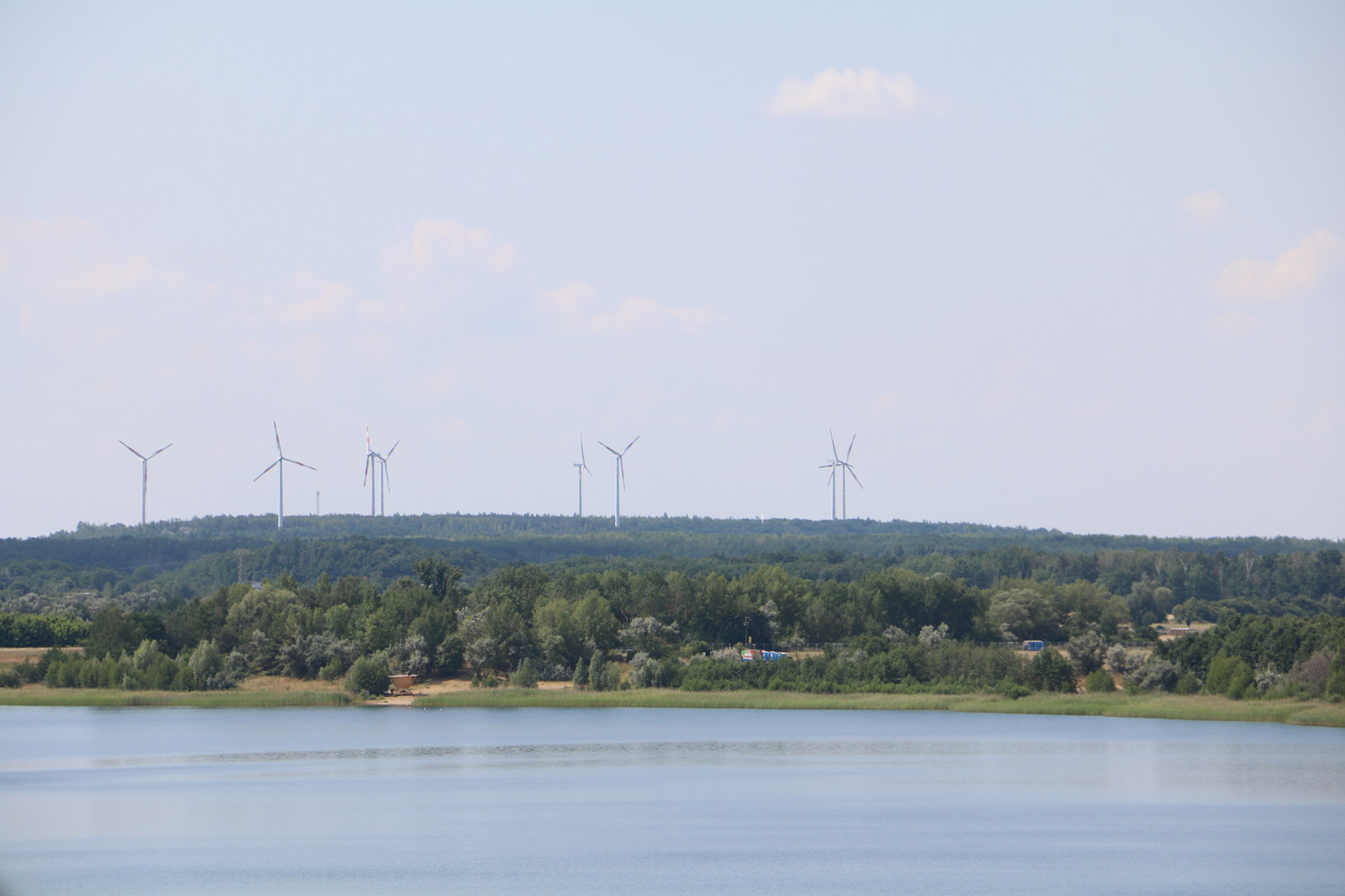 KI generiert: Das Bild zeigt eine Landschaft mit einem ruhigen See im Vordergrund und mehreren Windrädern in der Ferne auf einem bewaldeten Hügel. Die Szene vermittelt einen Eindruck von erneuerbarer Energie in einer natürlichen Umgebung.