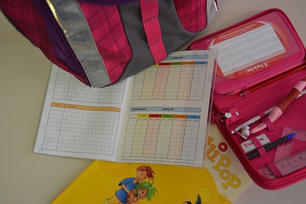 Hausaufgabenheft, Lehrbuch, Federmappe und Schulranzen stehen auf einem Tisch.