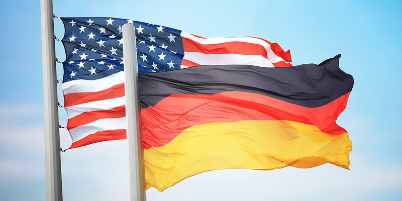 KI generiert: Das Bild zeigt die Flaggen der USA und Deutschlands, die im Wind wehen. Der blaue Himmel bildet den Hintergrund.