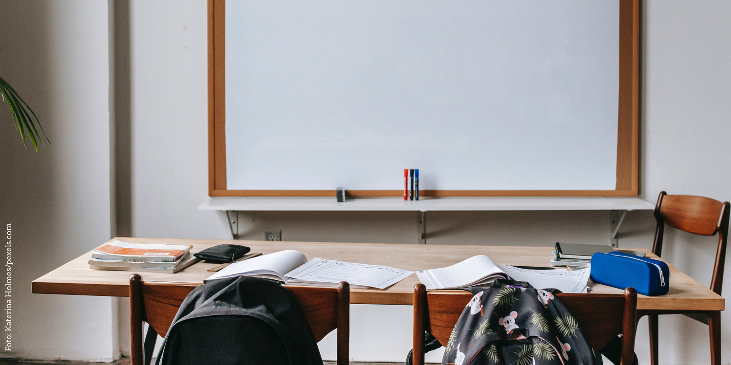 KI generiert: Das Bild zeigt einen leeren Klassenzimmerbereich mit einem Tisch, auf dem Bücher und Schulmaterial liegen. Im Hintergrund ist eine große, weiße Tafel zu sehen.