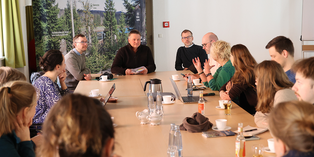 KI generiert: Das Bild zeigt eine Gruppe von Menschen, die um einen Tisch versammelt sind und offenbar eine Diskussion oder Besprechung führen. Auf dem Tisch befinden sich mehrere Getränke und Laptops, was auf ein formelles Treffen oder eine Konferenz hindeutet.