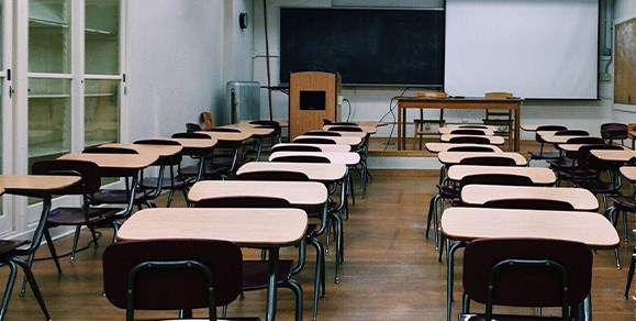 KI generiert: Das Bild zeigt ein leeres Klassenzimmer mit mehreren Reihen von Tischen und Stühlen sowie einem Tisch und einem Rednerpult vorne. Die Tafel und eine Leinwand sind ebenfalls sichtbar.