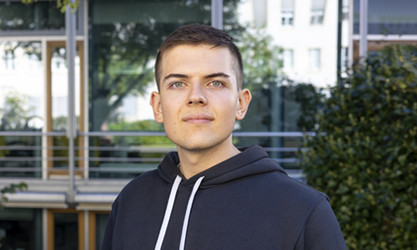 Alexander Sagandykov steht vor einer Gebäudestruktur und schaut in die Kamera.