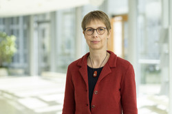 KI generiert: Das Bild zeigt eine Person mit Brille und einem roten Blazer, die in einem Innenraum mit viel natürlichem Licht steht. Der Raum wirkt modern und gut beleuchtet.