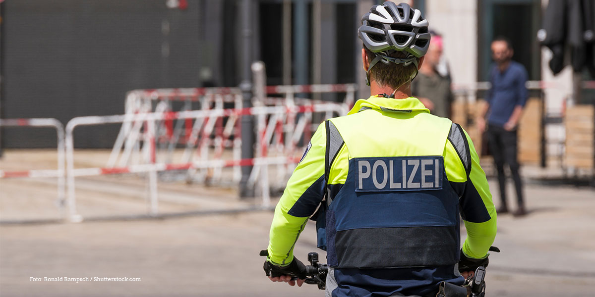 KI generiert: Das Bild zeigt einen Polizisten auf einem Fahrrad, der eine leuchtend gelbe Uniform und einen Helm trägt. Im Hintergrund sind Absperrungen und unscharfe Figuren zu erkennen.