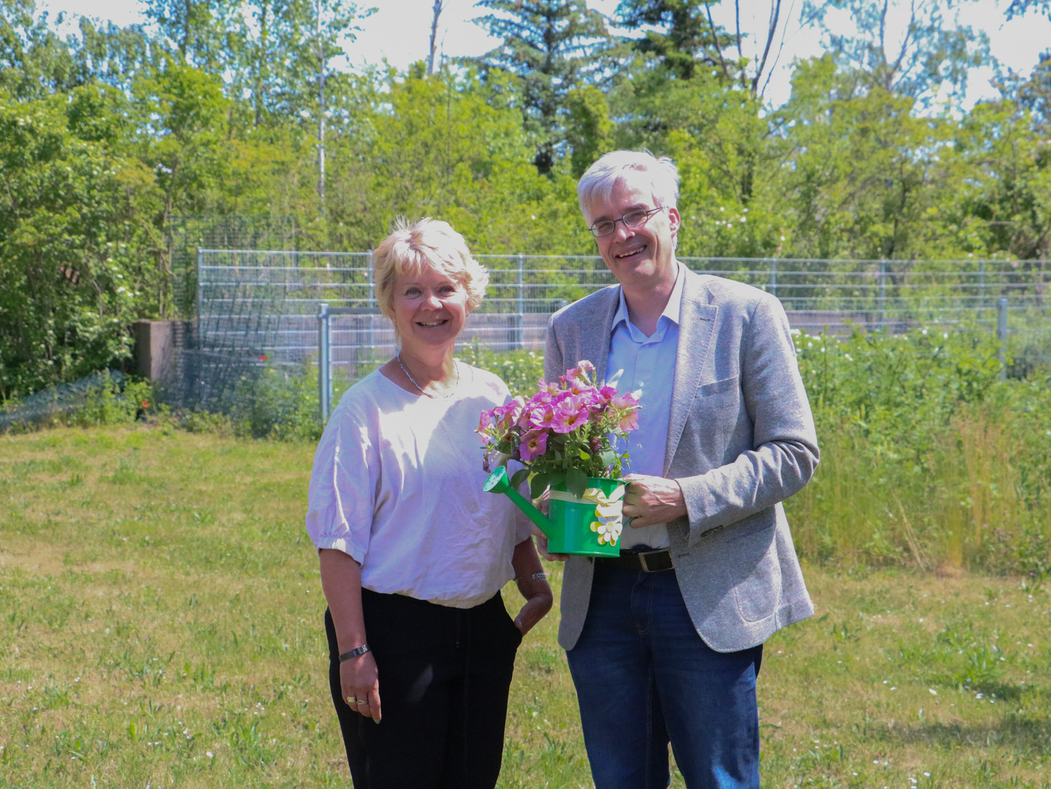 KI generiert: Ein Mann und eine Frau stehen in einem Garten oder Park. Der Mann hält einen grünen Eimer mit blühenden Blumen.