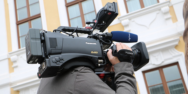 KI generiert: Das Bild zeigt eine Person, die eine professionelle Videokamera vor einem Gebäude bedient. Der Schwerpunkt liegt auf der Kameraausrüstung in einem urbanen Setting.