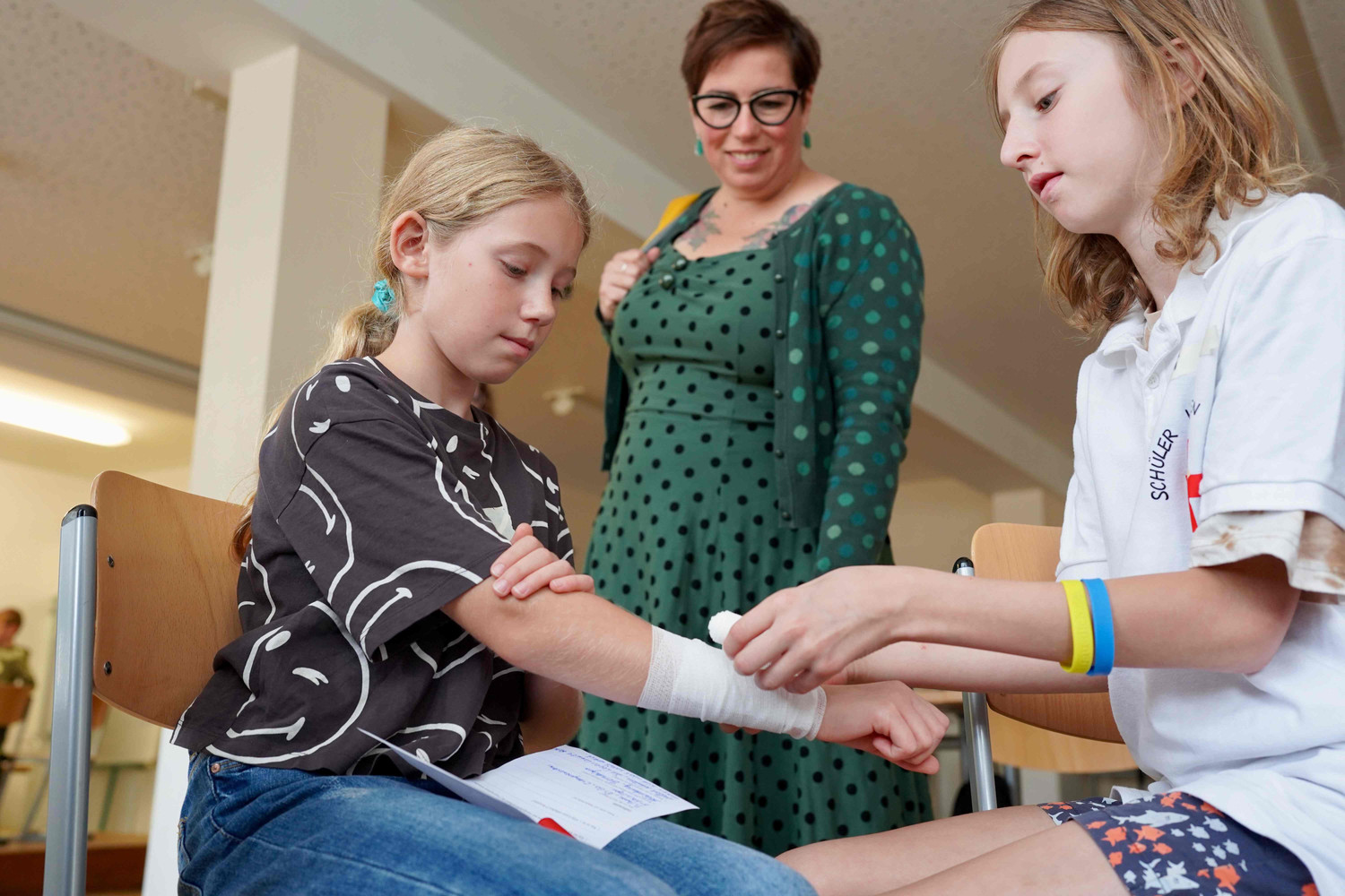 KI generiert: Zwei Kinder sind in einem Klassenzimmer zu sehen, wobei eines dem anderen einen Armverband anlegt. Eine erwachsene Frau steht daneben und beobachtet die Situation.