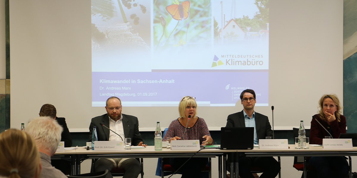 KI generiert: Das Bild zeigt eine Podiumsdiskussion zum Thema "Klimawandel in Sachsen-Anhalt" mit mehreren Teilnehmenden, die vor einer Präsentation sitzen. Der Fokus liegt auf dem Vortrag und denjenigen, die sprechen und diskutieren.