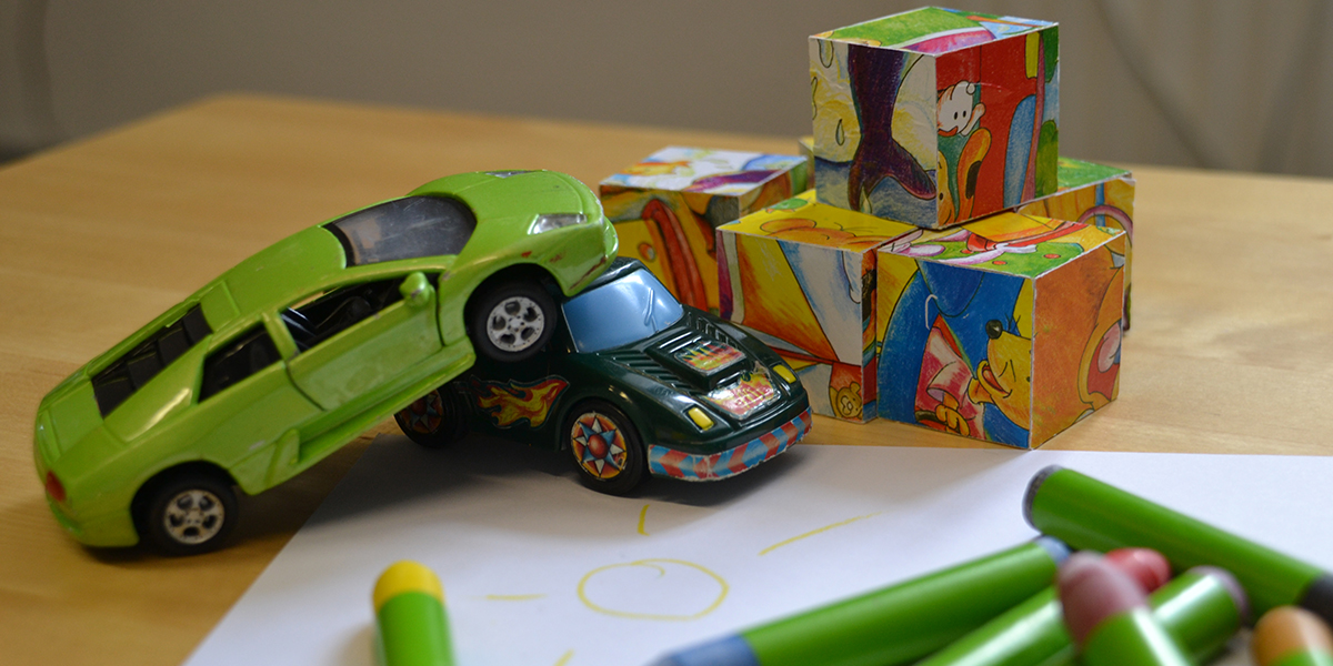 KI generiert: Das Bild zeigt zwei Spielzeugautos, die miteinander kollidiert sind, sowie bunte Bauklötze auf einem Tisch. Im Vordergrund sind Wachsmalstifte und ein Blatt Papier mit einer gezeichneten Sonne zu sehen.