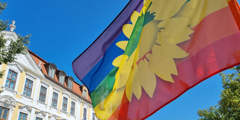 KI generiert: Das Bild zeigt eine Regenbogenflagge mit einem gelben Sonnenblumenmotiv vor einem historischen Gebäude und einem blauen Himmel. Der Hauptinhalt ist die Flagge, die im Wind weht.