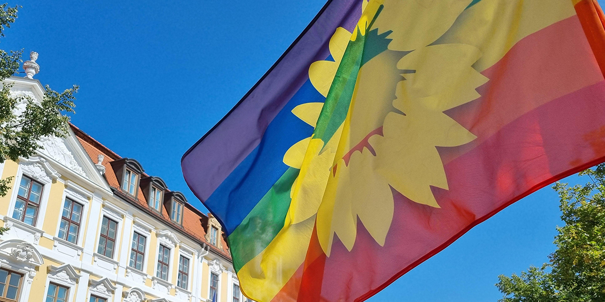 KI generiert: Das Bild zeigt eine Regenbogenflagge mit einem gelben Sonnenblumenmotiv vor einem historischen Gebäude und einem blauen Himmel. Der Hauptinhalt ist die Flagge, die im Wind weht.