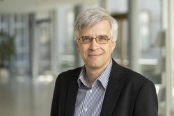 KI generiert: Das Bild zeigt einen Mann mit grauen Haaren und Brille in einem Anzug, der in einem modernen Innenraum lächelt. Der Fokus liegt auf seiner freundlichen, professionellen Ausstrahlung.