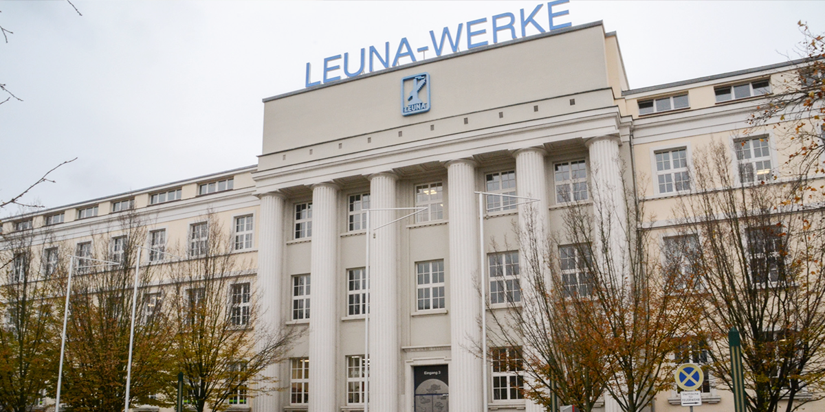 KI generiert: Das Bild zeigt das Hauptgebäude der LEUNA-WERKE mit seiner neoklassizistischen Architektur und der markanten Beschriftung auf dem Dach. Vor dem Gebäude sind kahle Bäume und ein Verkehrszeichen zu sehen.