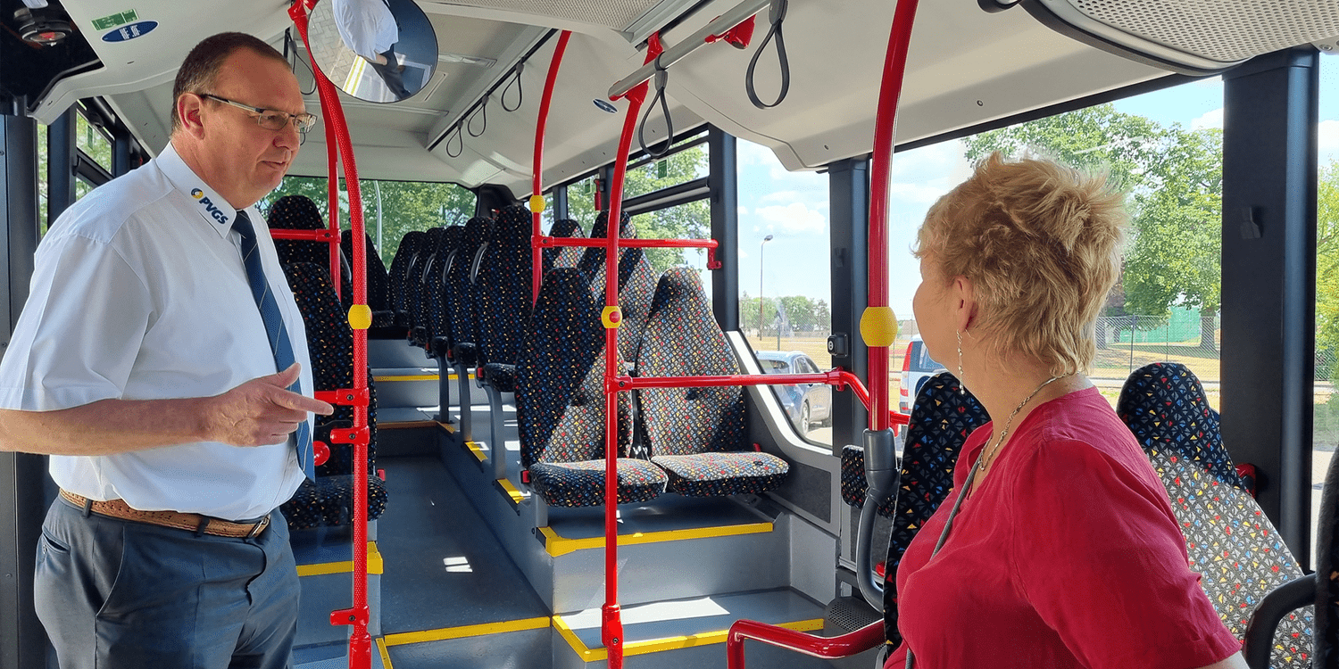 KI generiert: Auf dem Bild sieht man einen Mann in einem Busuniform vor einer Frau, die im Bus steht. Sie scheinen miteinander zu sprechen; der Bus ist leer und gut beleuchtet.