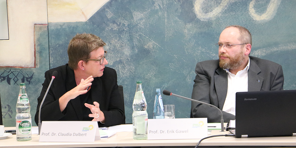 KI generiert: Das Bild zeigt zwei Personen, die an einem Tisch sitzen und miteinander diskutieren. Vor ihnen sind Namensschilder, Mikrofone und Wasserflaschen zu sehen.