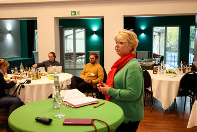 KI generiert: Das Bild zeigt eine Frau, die an einem Tisch mit Notizen steht, während im Hintergrund eine Gruppe von Menschen an einem runden Tisch sitzt. Es scheint sich um eine Besprechung oder Präsentation in einem Raum mit Tischen und Getränken zu handeln.
