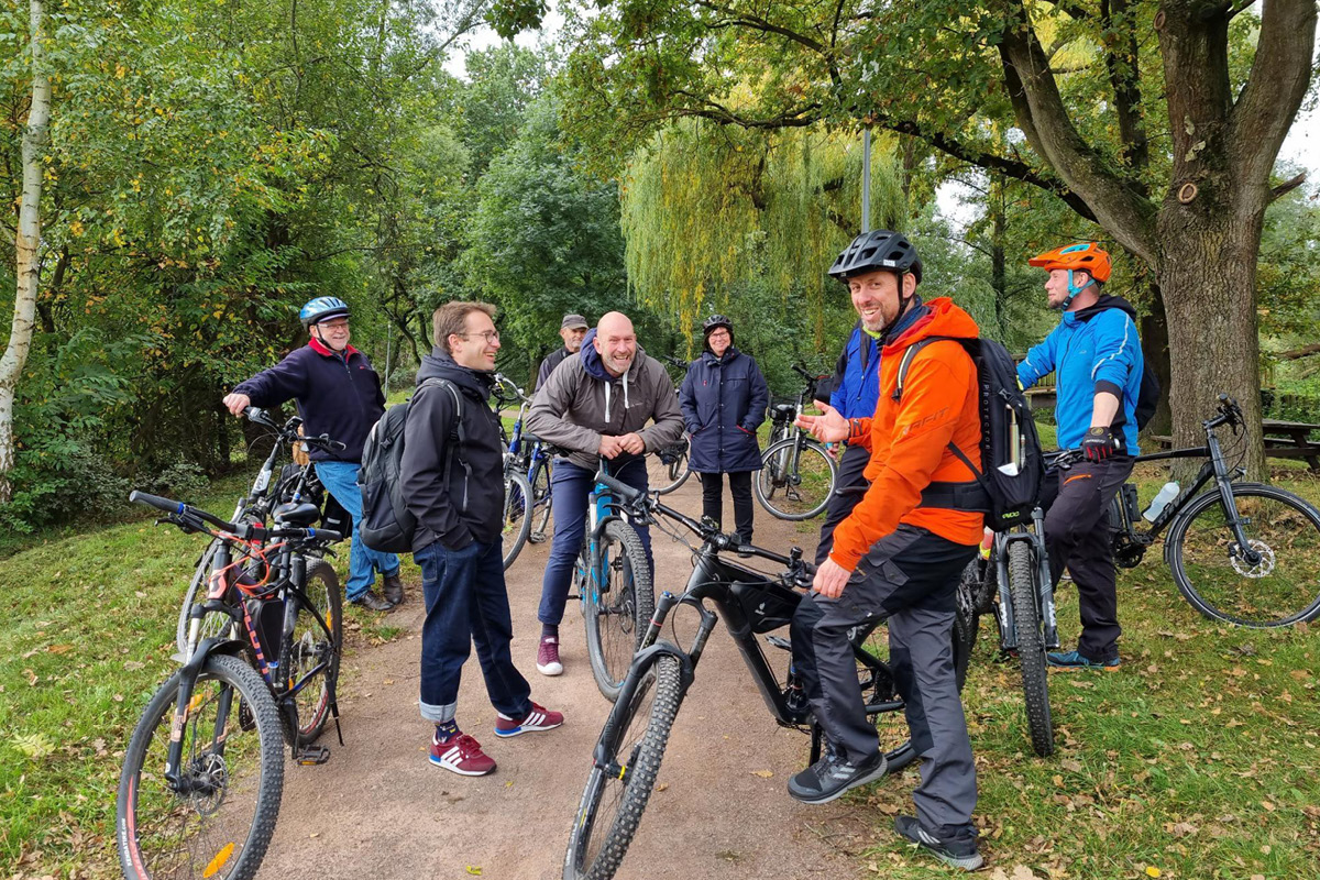 KI generiert: Eine Gruppe von Menschen mit Fahrrädern steht auf einem Waldweg und unterhält sich lächelnd. Sie tragen Freizeitkleidung und einige haben Fahrradhelme auf.