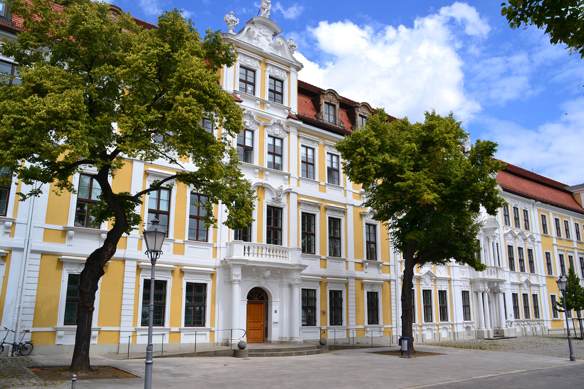 KI generiert: Das Bild zeigt ein historisches, mehrstöckiges Gebäude mit einer dekorativen Fassade und gelben Details. Vor dem Gebäude stehen mehrere Bäume, die den Gehweg säumen.