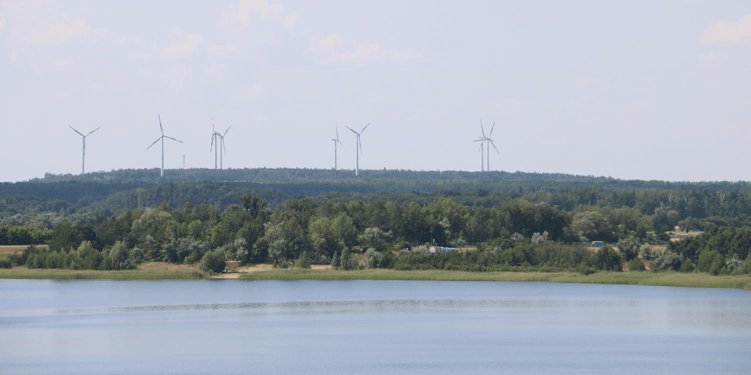 KI generiert: Das Bild zeigt eine Landschaft mit einem Gewässer im Vordergrund und einem Wald im Hintergrund, in dem mehrere Windkraftanlagen zu sehen sind. Der Hauptinhalt des Bildes sind die Windkraftanlagen auf einem bewaldeten Hügel.