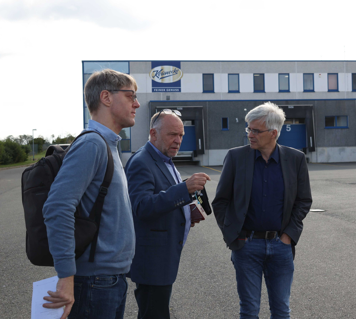 KI generiert: Das Bild zeigt drei Männer, die auf einem Parkplatz vor einem Gebäude miteinander sprechen. Im Hintergrund ist ein Gebäude mit dem Logo der Firma "Kowelek Feiner Genuss" sichtbar.