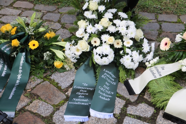 KI generiert: Das Bild zeigt Kränze mit weißen und gelben Blumen, die auf einem gepflasterten Boden liegen. Auf den Schleifen steht ein Gedenken an die Opfer des Nationalsozialismus.