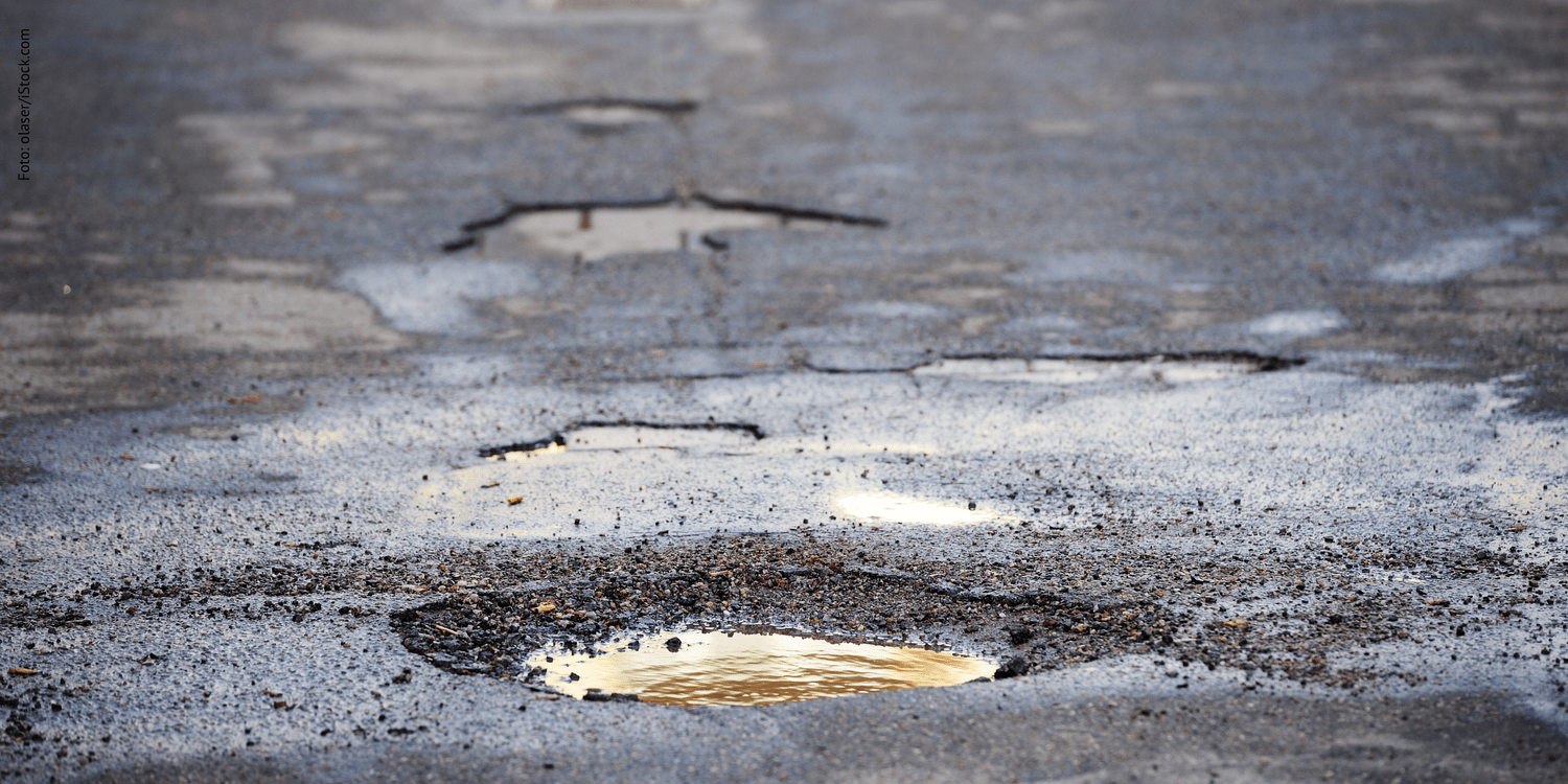 KI generiert: Das Bild zeigt eine beschädigte Asphaltstraße mit mehreren Schlaglöchern, die teilweise mit Wasser gefüllt sind. Das Hauptmotiv ist die unebene Oberfläche der Straße.