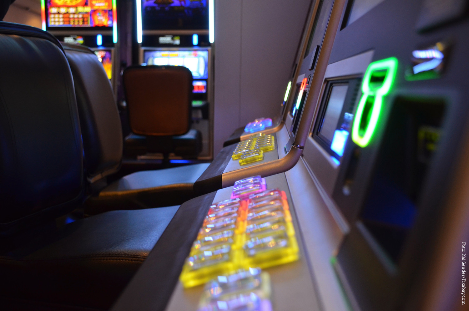KI generiert: Das Bild zeigt eine Reihe von Spielautomaten in einem Casino, mit leuchtenden Knöpfen und Bildschirmen. Im Vordergrund sind Sitzplätze vor den Automaten erkennbar.