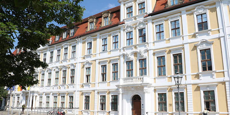 KI generiert: Das Bild zeigt ein historisches, mehrstöckiges Gebäude mit einer barocken Fassade. Es hat mehrere Fensterreihen und ein rotes Dach, und die Szenerie wird von einem Baum im Vordergrund ergänzt.
