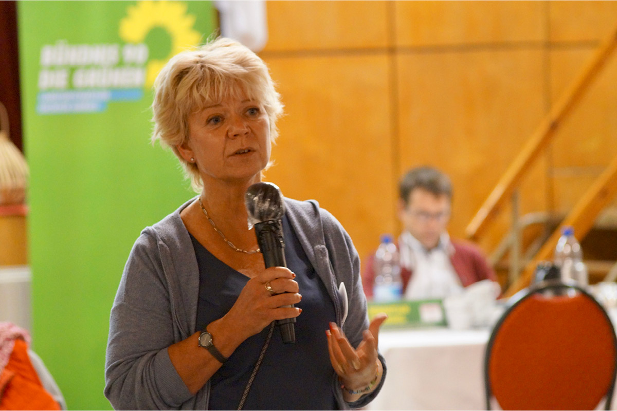 KI generiert: Die Hauptperson auf dem Bild spricht in ein Mikrofon, während im Hintergrund ein Banner mit dem Logo einer politischen Partei zu sehen ist. Es scheint sich um eine Veranstaltung oder Diskussion zu handeln.