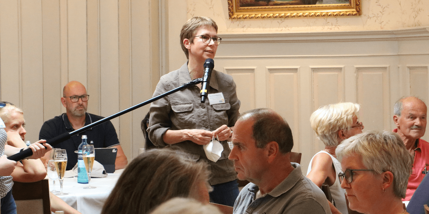 KI generiert: Das Bild zeigt eine Frau, die während einer Veranstaltung in einem Raum steht und in ein Mikrofon spricht, während Menschen ihr aufmerksam zuhören. Die Szene wirkt wie ein gesellschaftliches Treffen oder eine Diskussionsrunde.