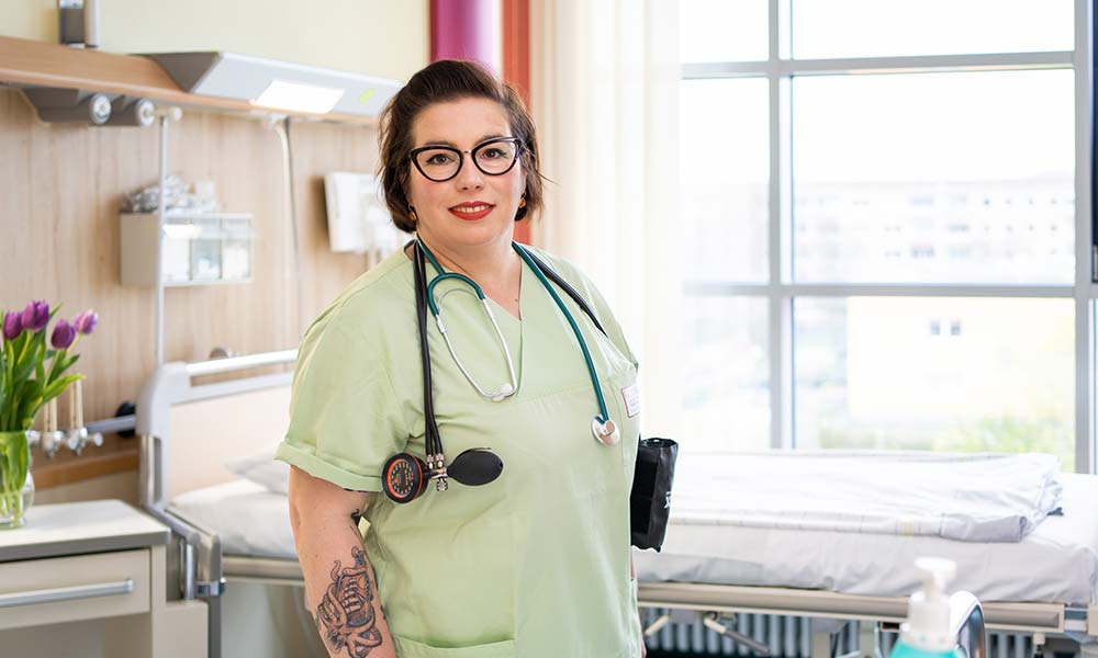 KI generiert: Das Bild zeigt eine Krankenschwester in einem Krankenhauszimmer, die mit einem Stethoskop und medizinischer Kleidung ausgestattet ist. Im Hintergrund sind ein Krankenbett und ein Fenster zu sehen.