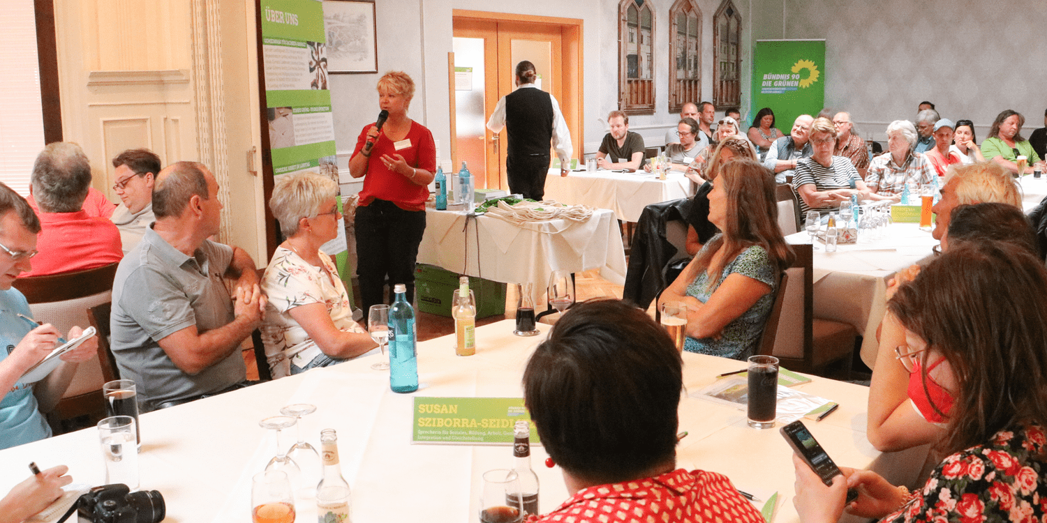 KI generiert: Das Bild zeigt eine Versammlung in einem Raum, bei der eine Frau mit einem Mikrofon zu einer Gruppe von Menschen spricht, die an Tischen sitzen. Im Hintergrund sind Banner der Partei Bündnis 90/Die Grünen zu sehen.