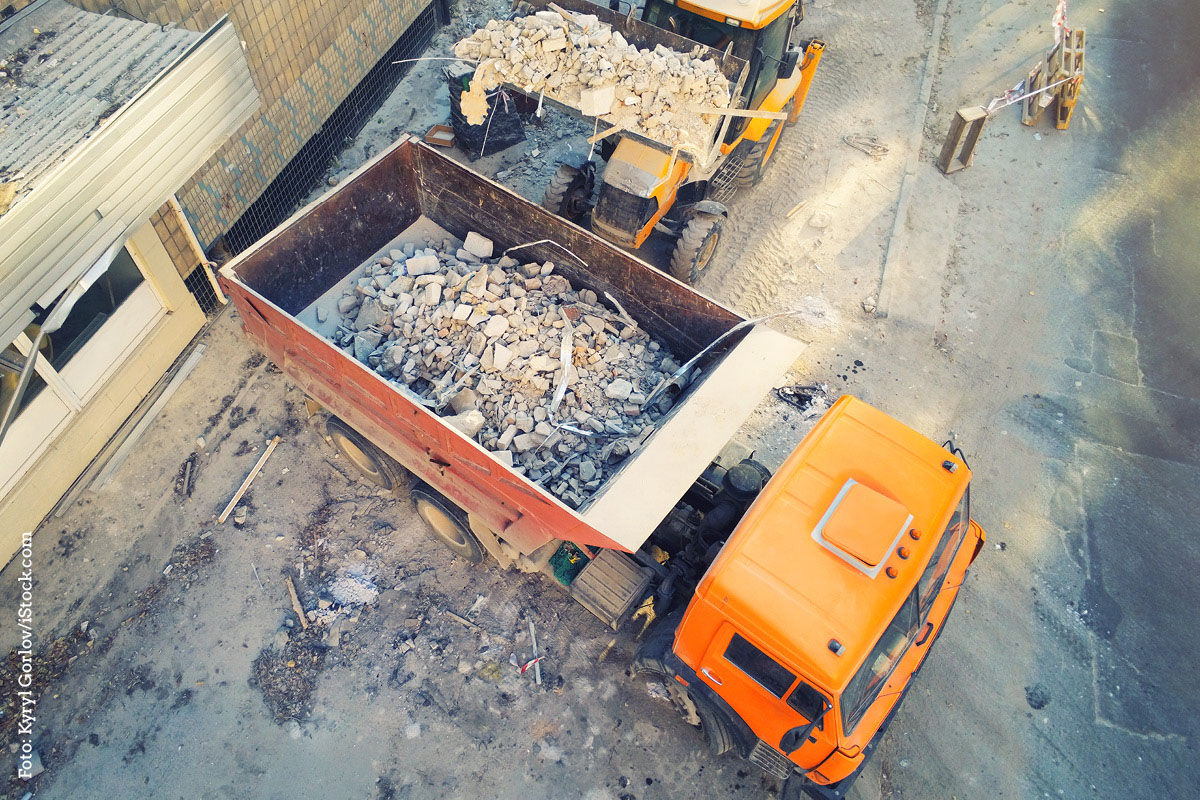 KI generiert: Das Bild zeigt eine Baustellenszene mit einem Bagger, der Schutt in einen orangefarbenen LKW lädt. Die Umgebung ist staubig, und es liegt viel Bauschutt herum.