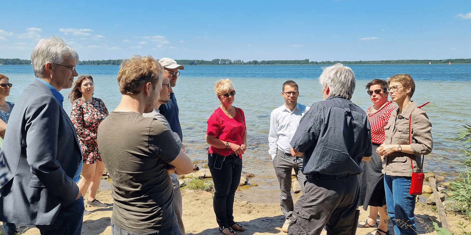 KI generiert: Eine Gruppe von Menschen steht an einem Ufer und scheint in ein Gespräch vertieft zu sein. Im Hintergrund ist ein ruhiger See unter einem klaren blauen Himmel zu sehen.
